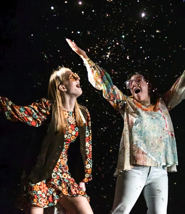 Two performers on stage in colorful clothing, throwing glitter with mouths open wide.