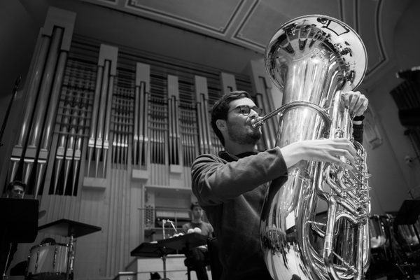 A tuba player, viewed from below, plays in front of a large organ.