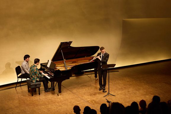 Two pianists sit at a piano keyboard in concert.