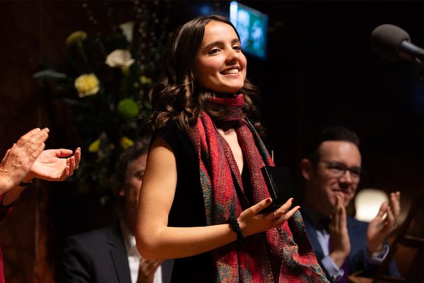 Sofía Patterson-Gutiérrez stands on a stage holding a black metal trophy. She is wearing a black dress and red scarf. Members of the audience can be seen clapping.
