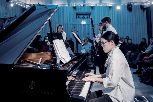 A piano student can be seen playing, alongside other musicians. An audience is sat in the background of a blue lit room.