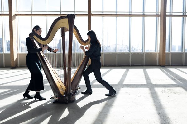 Two musicians are playing the harp. They are wearing all black. They stand in an empty room against large windows, with the London landscape in the background.