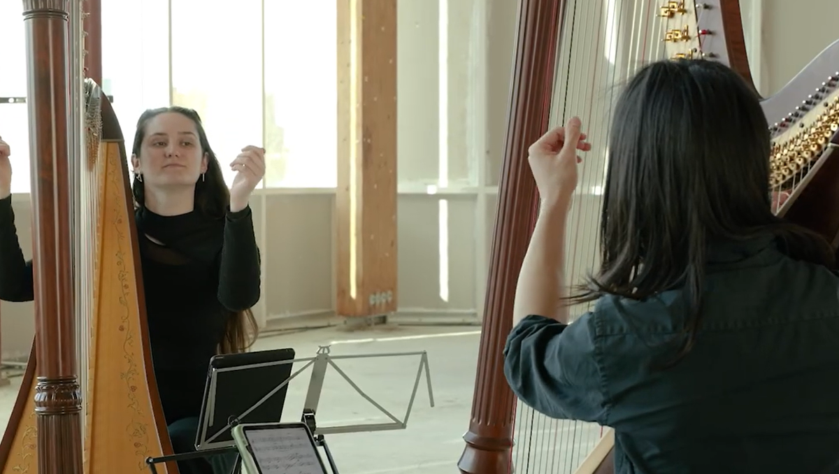 Two harpists sit at their harps. Their hands are held up next to their harps.