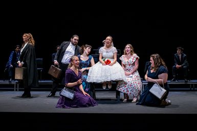 Performers on stage. A bride is sat centre, crying, with guests surrounding her singing.