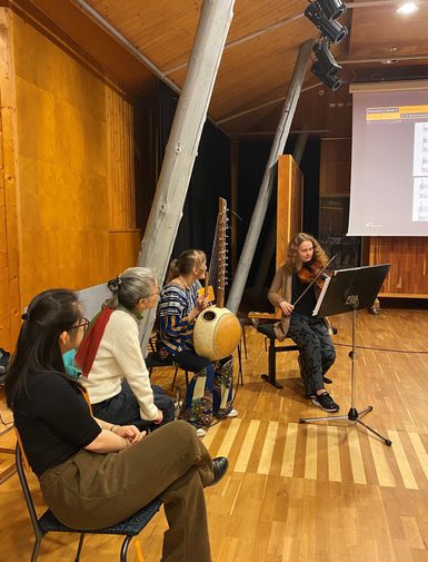 Four musicians sit in a semicircle in a large wooden practise room. A violinist plays as the others watch.