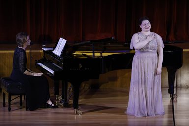 A singer wearing a pale pink dress stands on stage, with her hand against her shoulder. She is joined by a pianist in a black dress.