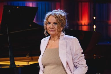 A woman stands in front of a piano, wearing a beige top and purple blazer