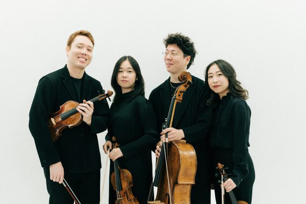 Four musicians stand against a white background. They are posing for a picture holding string instruments