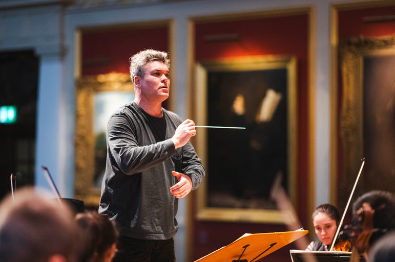 A conductor wearing casual clothes conducts on stage. Portraits can be seen in the background.