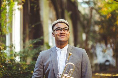Aaron Azunda Akugbo stands against a woodland background holding a trumpet