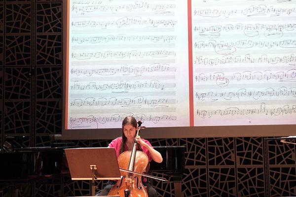 A musician plays the cello on stage, in front of a large projection of sheet music