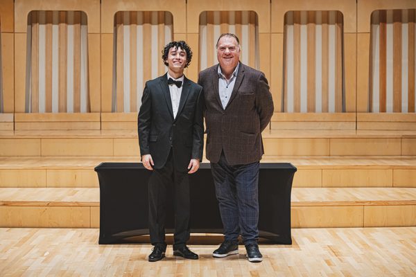 David Karapetian stands on stage, in front of a black table, with Sir Bryn