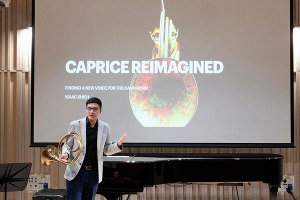 Man standing in front of a presentation slide and piano holding a french horn