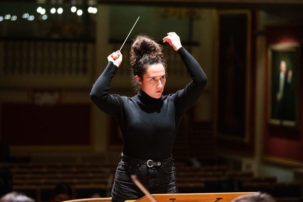 A female conductor waves a baton. She is stood on stage.