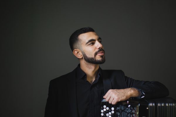 Portrait image of Flavio Rodrigues looking off to the right, leaning his hand on an accordion.