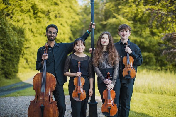 Isla Quartet stand holding their string instruments. One member of the group holds a lamppost. Grass and trees can be seen in the background.