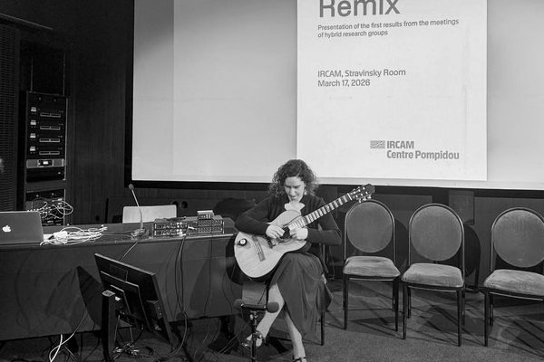 Katalin Koltai sits on stage holding a guitar. She sits in front of a large screen, seats and a desk