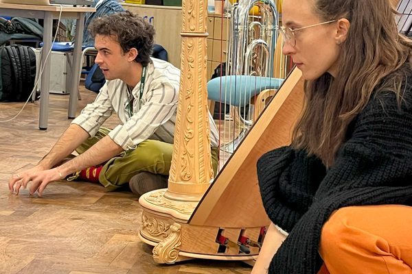 Two musicians sit or kneel on the floor of a classroom, next to a harp