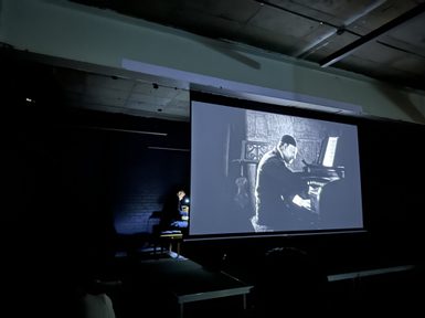A man playing a keyboard in a dark space behind a screen showing an old black and white film of a man playing a keyboard.