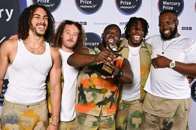 Ezra Collective stand together on the Mercury Prize red carpet. One member of the group holds an award towards the camera.