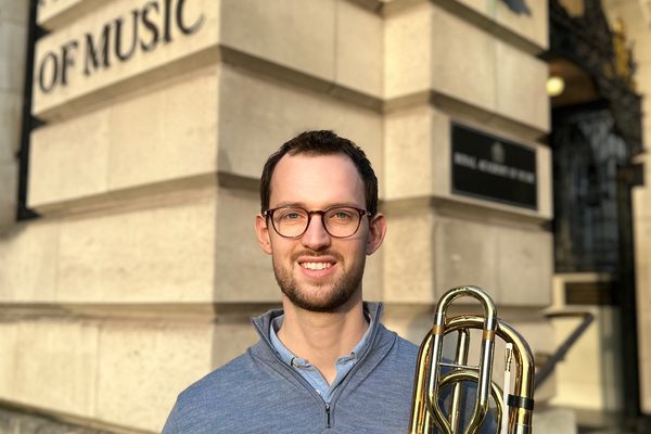 James Buckle, standing outside the Royal Academy of Music, smiling, and holding a bass trombone