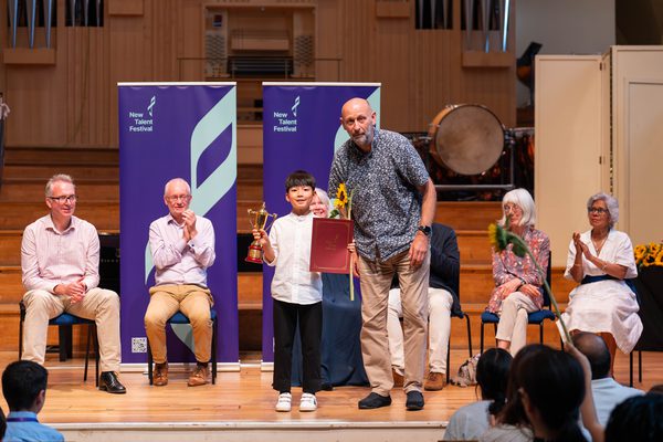 A young musician being presented an award on a stage, with several adults sat behind them applauding