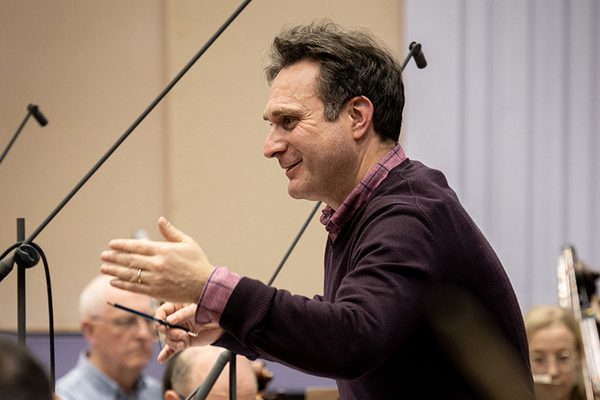 Richard Tanner stands gesturing. Musicians can be seen in the background.