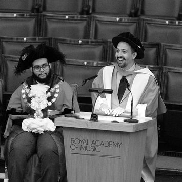 Lin Manuel Miranda stands at a lecturn in graduation outfit, smiling.