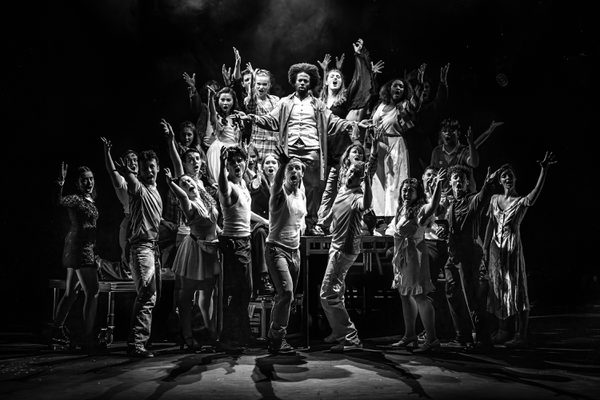 A large musical theatre cast huddle together in a group and sing towards the audience.