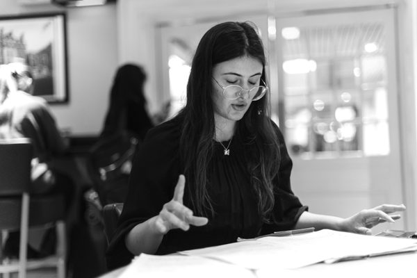 A woman wearing glasses and a black shirt sits at a table, reviewing papers spread out in front of her.