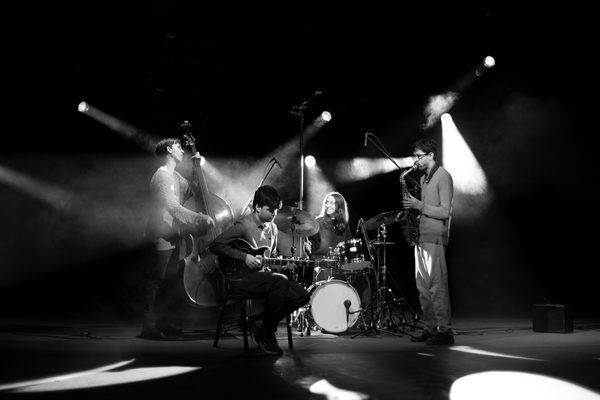 A band of four musicians performs in black and white, lit by spotlights and a smokey background.