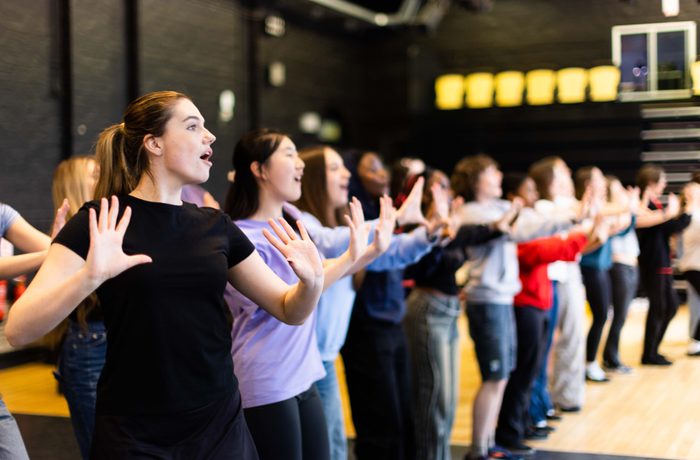A row of young performers in rehearsal, raising their hands and singing.
