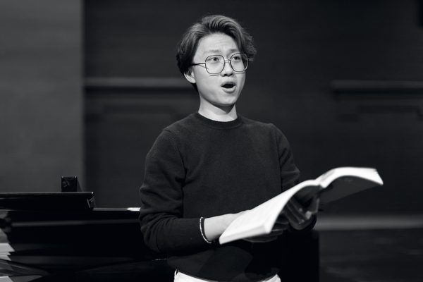 A vocal studies student holds sheet music in hand while looking out of frame, singing (black and white).