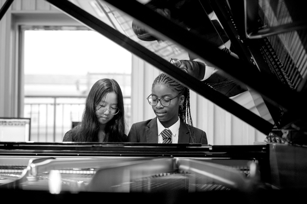 Two young students play at a piano keyboard, smiling.
