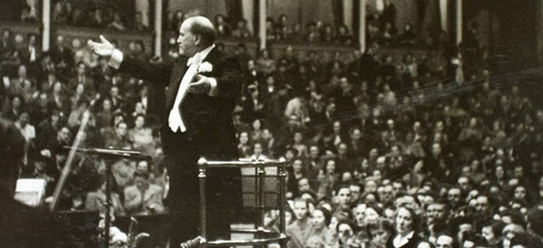 Scanned black and white photo of Sir Henry Wood stood atop a conductors podium, with raised arms in front of a busy concert audience.