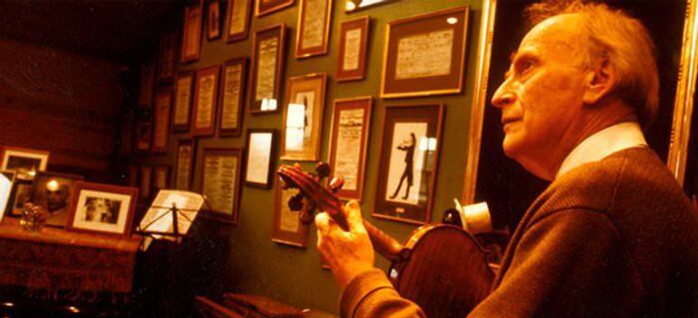 A violin player viewed from behind their left shoulder, plucking the instrument in front of a wall of framed certificates.