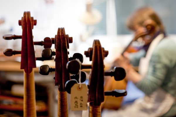 A person plays a violin out of focus in the background, with a row of violins on a rack in the foreground.