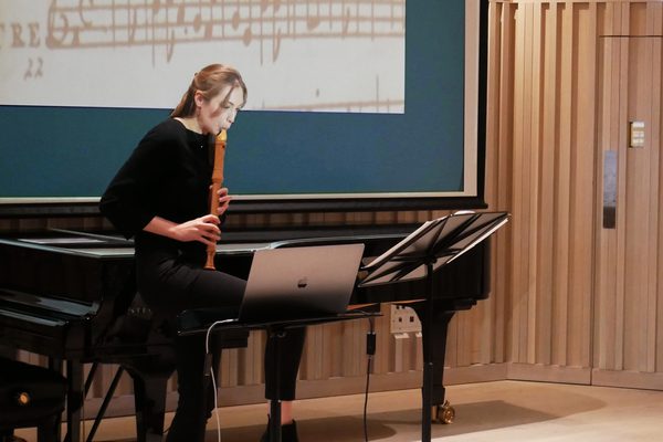 A research student performs to an audience reading sheet music, standing on one leg to achieve a high note.