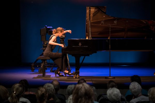 A woman on stage playing a grand piano for a concert audience, under a blue spotlight.