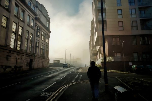 A person in shadow walking between two tall buildings separated by a city road with a misty cloud between them in the distance.