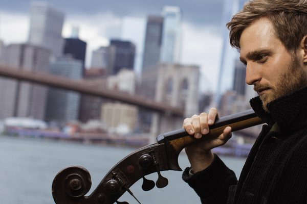 A side profile of a man holding a double bass over his shoulder, the scroll and pegs in front of him, with a city river background.