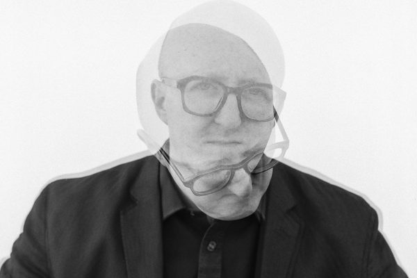 Black and white headshot of a man facing the camera wearing dark framed glasses with a superimposed headshot looking downwards.