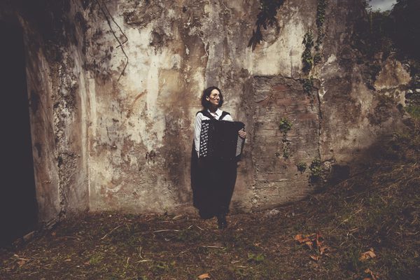A woman standing in front of an old distressed plaster wall holding an accordion.