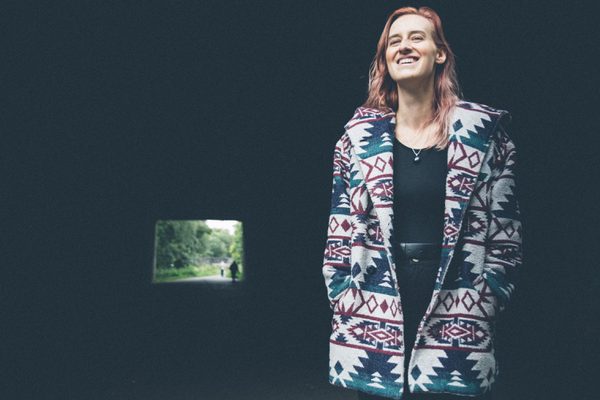 A woman standing smiling in a brightly coloured patterned coat against a dark background, with a small square of light behind from a tunnel opening.