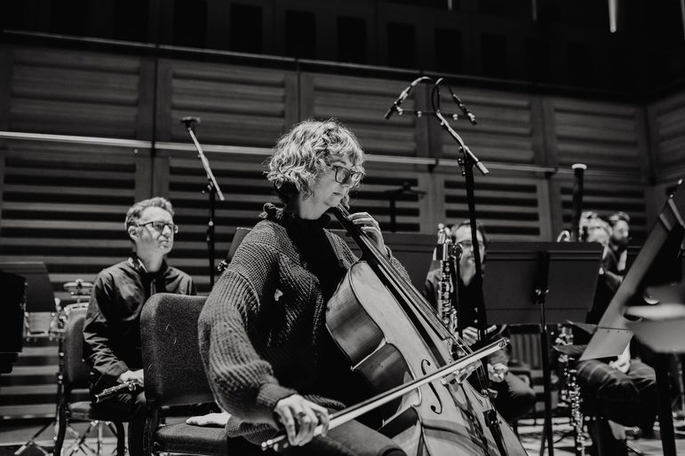Black and white image of a woman seated with a cello, on stage as part of an ensemble.
