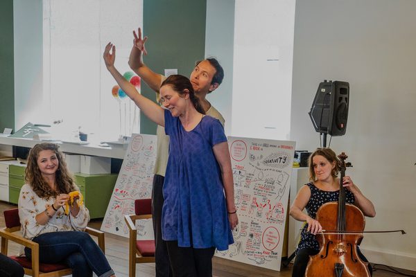 A group of seated and standing workshop participants smile and make expressive hand gestures, accompanied by a cellist.