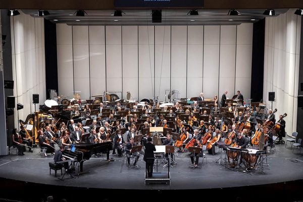An orchestra sits on a stage with a white backdrop.