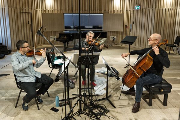 Three musicians sitting together in a semi-circle in a studio playing violin, viola and cello, with microphones and cables to record their sound.