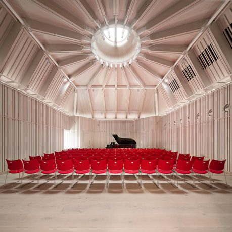 A recital hall with wood panelling, red chairs and a grand piano.
