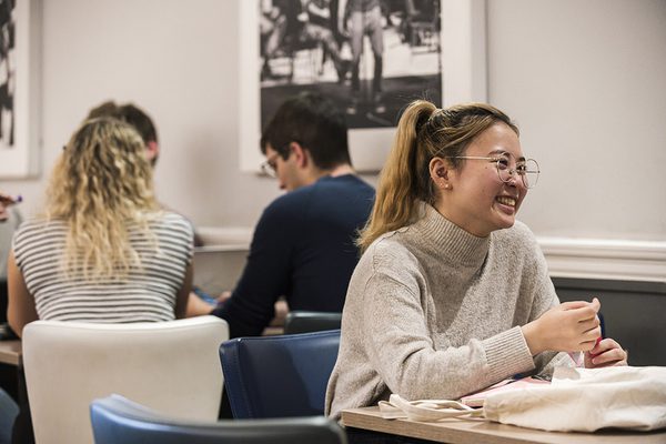 A students sits in the cafeteria at a table, smiling to something off camera.
